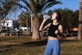 Active young fit woman in wireless headphones drinks water from bottle after exercising outdoors at palm trees park Royalty Free Stock Photo