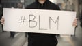 Activist wearing dark clothes holds a placard with BLM or Black Lives Matter hashtag