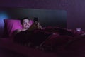 Addicted guy play with the smart phone at night in the bed. Man with beard lying down in hotel room typing email and smiling.