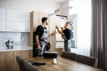 Adding care to your technologies. Aged repairman in uniform fixing refrigerator in the kitchen, while his colleague