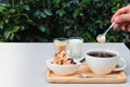 Adding sugar to coffee. Cup of coffee, cookie, milk and sugar are on wood tray on white table in the garden atmosphere. Royalty Free Stock Photo