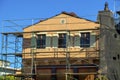 Adobe style house design with shutters and double gable roof under construction with metal scafolding in sun blue sky