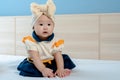 Adolable Asian newborn baby girl, big eyes, puffy cheeks, cutely dressed in yellow and blue, sitting on a white bed looking at Royalty Free Stock Photo