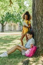 African american schoolgirl standing near friend sitting on lawn under tree and reading book Royalty Free Stock Photo