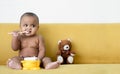 Adorable African baby newborn in diaper sitting on sofa with small bear doll trying to grab a spoon to feed herself. Royalty Free Stock Photo