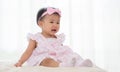 Adorable Asian baby crying, sitting on bed at home. Unhappy little cute toddler girl wear casual pink dress, headband and socks. Royalty Free Stock Photo