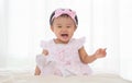 Adorable Asian baby laughing, looking at camera, sitting on bed. Smiling little cute toddler girl wear casual pink dress, headband Royalty Free Stock Photo