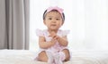 Adorable Asian baby smiling, clapping hands, sitting on bed. Little cute toddler girl wear casual pink dress, headband and socks. Royalty Free Stock Photo