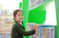 Adorable Asian little girl looking for book on bookshelf at library. Child reaches for books on a shelf Royalty Free Stock Photo
