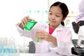 Adorable Asian schoolgirl in lab coat doing science experiments, pouring green reagent solution from beaker into test tube Royalty Free Stock Photo