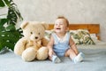 Adorable baby boy sitting on the bed and laughs in white sunny bedroom with toy teddy bear and green plant monstera. Newborn child Royalty Free Stock Photo