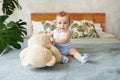 Adorable baby boy in white sunny bedroom holding toy teddy bear with green plant monstera. Newborn child relaxing in bed. Textile Royalty Free Stock Photo