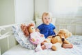 Adorable baby girl in blue dress sitting on bed and playing with doll and teddy bear Royalty Free Stock Photo