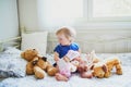 Adorable baby girl in blue dress sitting on bed and playing with doll, teddy bear and dog Royalty Free Stock Photo