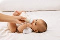 Adorable baby girl, child, in white onesie lying on bed and eating from bottle against white background. Nutrition Royalty Free Stock Photo