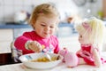 Adorable baby girl eating from fork vegetables and pasta. Little child feeding and playing with toy doll. Cute toddler