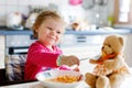 Adorable baby girl eating from fork vegetables and pasta. Little child feeding and playing with toy teddy bear. Royalty Free Stock Photo