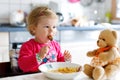 Adorable baby girl eating from fork vegetables and pasta. Little child feeding and playing with toy teddy bear. Royalty Free Stock Photo