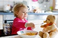 Adorable baby girl eating from fork vegetables and pasta. Little child feeding and playing with toy teddy bear. Royalty Free Stock Photo