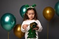 Cute baby girl in elf hoop holds her gift in green glitter wrapping paper and golden ribbon, stands against gray background with Royalty Free Stock Photo