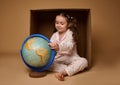 Adorable baby girl in pajamas sitting ahead a cardboard box and examining the Earth globe,  over beige background with Royalty Free Stock Photo