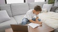 Adorable blond boy, a budding wordsmith, engrossed in putting pen to paper with his laptop\'s assistance, writing his ideas in a Royalty Free Stock Photo