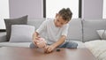 Adorable blond boy, financial whizz-kid, relaxing on sofa, comfortably emptying piggy bank as part of indoor savings activity at