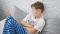 Adorable blond boy in pyjamas, sitting on bed in his bedroom, hugging teddy bear with sad expression, showing signs of unhappiness Royalty Free Stock Photo