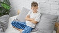 Adorable blond boy, sitting on bed with arms crossed, looking upset in bedroom - a sad morning scenario Royalty Free Stock Photo