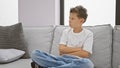 Adorable blond boy sitting in a cozy living room, arms crossed in an upset expression, exuding a serious vibe on the comfortable Royalty Free Stock Photo