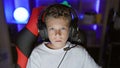 Adorable blond boy streamer, serious face focused on game, playing in dark gaming room with digital tech, sporting headset and Royalty Free Stock Photo