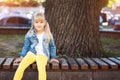 Adorable blond caucasian preschooler fashionista girl wearing jeans and bright yellow leggins sitting over wooden bench and