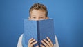 Adorable blonde boy student, mouth covered with book, standing isolated on a blue background, diving into learning with passion