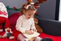 Adorable blonde girl reading book sitting on sofa by christmas decor at home Royalty Free Stock Photo