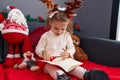 Adorable blonde girl reading book sitting on sofa by christmas decor at home Royalty Free Stock Photo