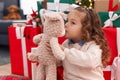 Adorable blonde toddler kissing teddy bear sitting on floor by christmas tree at home