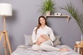 Adorable calm pregnant brown haired caucasian woman sitting in bed resting embracing her belly looking at camera resting at home