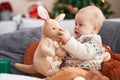 Adorable caucasian baby holding kangaroo doll sitting on sofa by christmas tree at home Royalty Free Stock Photo