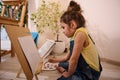 Adorable Caucasian child, cute little girl sitting on the floor at a wooden easel and drawing picture painting on canvas. Art Royalty Free Stock Photo