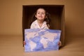 Adorable Caucasian child 4 years old girl sitting inside a cardboard box with a poster- painted image with World map, 