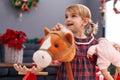 Adorable caucasian girl holding baby doll and horse toy standing by christmas decoration at home Royalty Free Stock Photo
