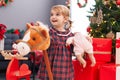 Adorable caucasian girl holding baby doll and horse toy standing by christmas tree at home Royalty Free Stock Photo