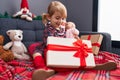 Adorable caucasian girl holding gift and baby doll sitting on sofa by christmas decoration at home Royalty Free Stock Photo