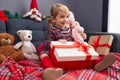 Adorable caucasian girl holding gift and baby doll sitting on sofa by christmas decoration at home Royalty Free Stock Photo