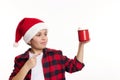 Handsome preteen boy in Santa hat, pointing on a red cup of hot cocoa with marshmallows, isolated on white background Royalty Free Stock Photo