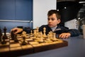 Adorable smart Caucasian child boy sitting at a table and playing chess, moving a chess piece on a chessboard