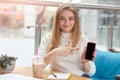 Adorable cheerful young Caucasian woman holds smart phone and points fore finger at black display, showing copyspace  over Royalty Free Stock Photo