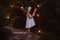 Adorable child girl in white dress with book in summer evening garden decorated with lights