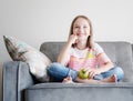 Adorable child happy girl 8 years old sits on a gray sofa with a green apple in hands Royalty Free Stock Photo