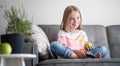 Adorable child girl 8 years old sits on a gray sofa with a green apple and a TV remote control in her hands rest and entertainment Royalty Free Stock Photo